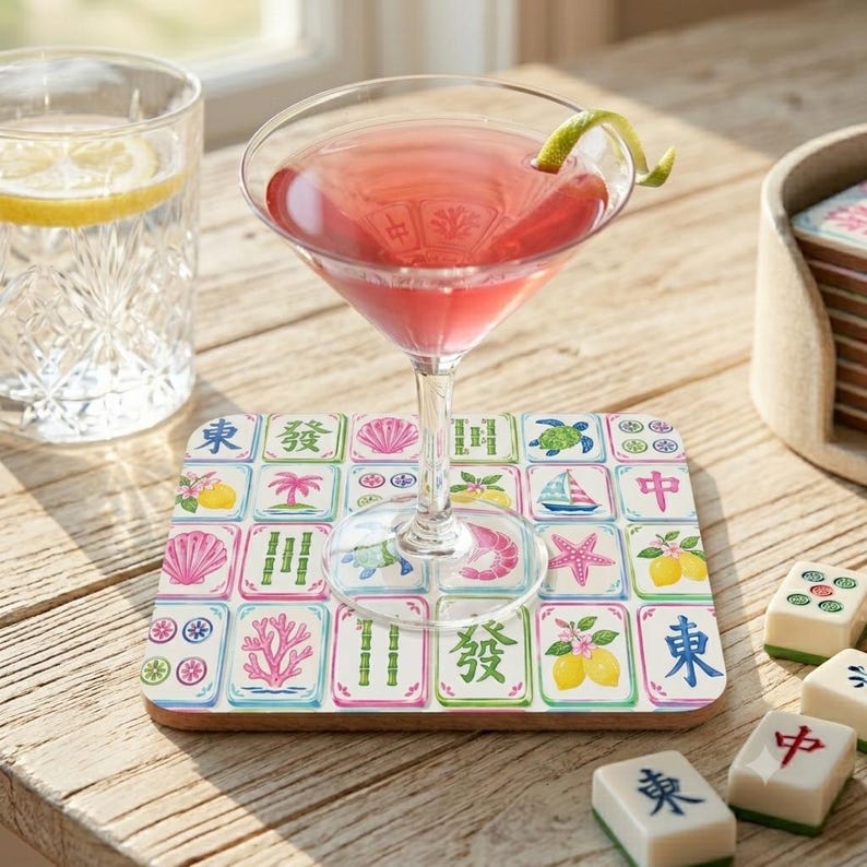 May include: A square coaster featuring a grid of colorful illustrations inspired by mahjong tiles, including pink coral, green bamboo, yellow lemons, and blue sailboats. A martini glass with a pink cocktail and a lime twist sits on top.