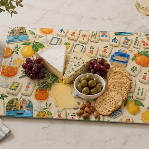 May include: A rectangular serving board with a colorful design featuring Mahjong tiles, oranges, and cocktails. The board holds cheese, grapes, olives, crackers, and almonds. A glass of white wine and a bottle of wine are in the background.