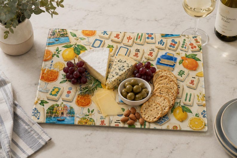 May include: A rectangular serving board with a colorful design featuring Mahjong tiles, oranges, and cocktails. The board holds cheese, grapes, olives, crackers, and almonds. A glass of white wine and a bottle of wine are in the background.