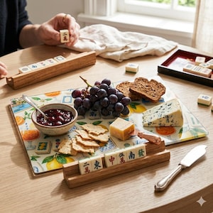 May include: A wooden Mahjong tile holder with tiles, a serving tray with food, and a bowl of jam. The tray includes grapes, cheese, crackers, and bread. A person is holding a tile. The scene is set on a wooden table.