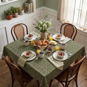 May include: A square dining table covered with a sage green tablecloth featuring a woven lattice pattern. The table is set with four white bowls of oatmeal topped with berries, four mugs, glasses of orange juice, and a basket of bread.