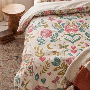 May include: A cream-colored duvet cover and matching pillowcase featuring a repeating pattern of pink roses, teal leaves, yellow stars, crescent moons, butterflies, hearts, and decorated elephants in pink, teal, and yellow hues.