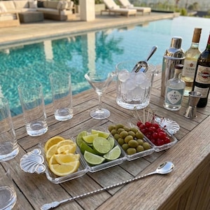 May include: A bar setup with cocktail ingredients and tools. Includes a clear ice bucket, cocktail shaker, bottles of liquor, a tray with lemon, lime, olives, and cherries, and a long bar spoon. The scene is set on a wooden table near a pool.