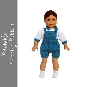 May include: A knitted doll outfit with a teal blue overall and a white long-sleeved shirt. The doll is wearing white socks.
