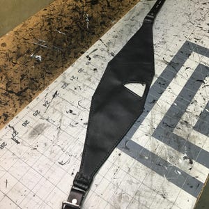 May include: A black leather harness with a buckle and a cut-out design. The harness is lying on a gray and white checkered surface.