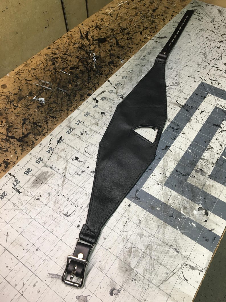 Peut inclure: Un harnais en cuir noir avec une boucle et un motif d&eacute;coup&eacute;. Le harnais est pos&eacute; sur une surface &agrave; carreaux gris et blancs.