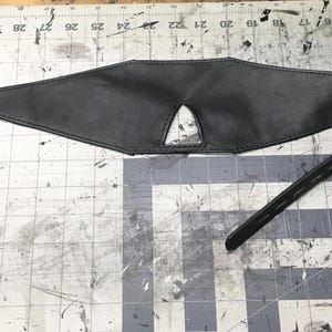 Peut inclure: Un bandeau en cuir noir avec un petit trou pour les yeux au centre. Le bandeau est attach&eacute; &agrave; une longue sangle avec une boucle en m&eacute;tal.