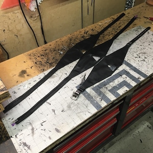 Peut inclure: Trois sangles en cuir noir avec des boucles m&eacute;talliques, dispos&eacute;es sur une surface quadrill&eacute;e blanche avec des marques noires. Les sangles sont dispos&eacute;es en forme de triangle, les pointes du triangle &eacute;tant orient&eacute;es vers le bas de l'image.