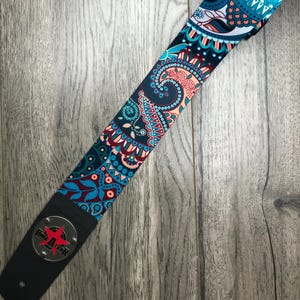 May include: A guitar strap featuring a vibrant paisley pattern in shades of teal, dark blue, and coral. The strap includes a black leather end piece with a circular silver metal plate displaying a red star and the text Rock Star in black letters.