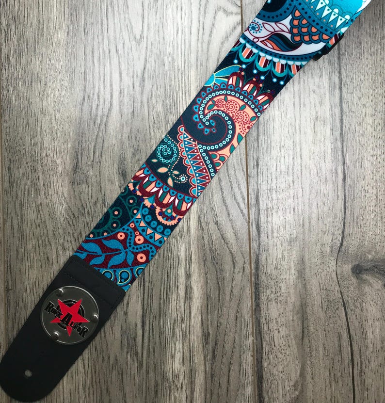 May include: A guitar strap featuring a vibrant paisley pattern in shades of teal, dark blue, and coral. The strap includes a black leather end piece with a circular silver metal plate displaying a red star and the text Rock Star in black letters.