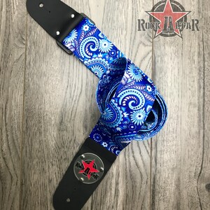 Pode incluir: Uma correia de guitarra com um padr&atilde;o floral paisley vibrante em azul e branco. A correia inclui extremidades em couro preto com ferragens prateadas e um emblema circular de metal com uma estrela vermelha e o texto Rock Altar em preto.
