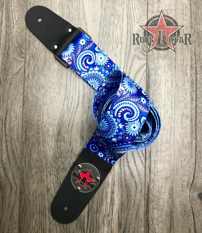 Pode incluir: Uma correia de guitarra com um padr&atilde;o floral paisley vibrante em azul e branco. A correia inclui extremidades em couro preto com ferragens prateadas e um emblema circular de metal com uma estrela vermelha e o texto Rock Altar em preto.