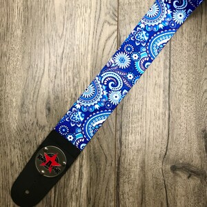 Pode incluir: Uma correia de guitarra com um padr&atilde;o floral e paisley vibrante em azul, branco e azul claro. A correia inclui uma extremidade de couro preto com uma placa circular de metal prateado exibindo uma estrela vermelha e o texto Rock Star em preto.