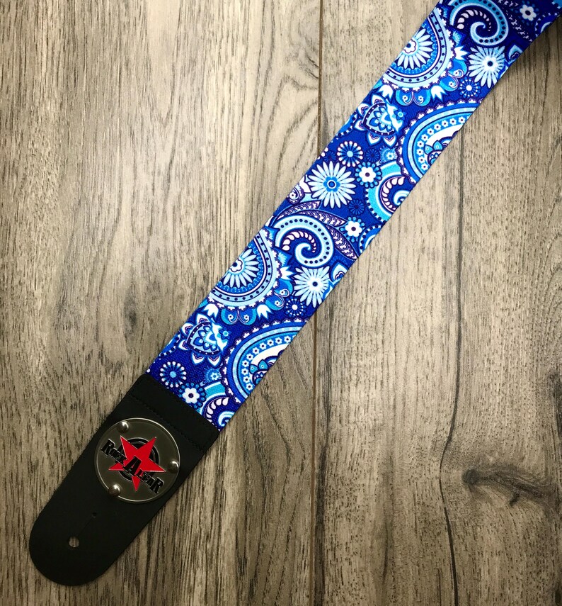 Pode incluir: Uma correia de guitarra com um padr&atilde;o floral e paisley vibrante em azul, branco e azul claro. A correia inclui uma extremidade de couro preto com uma placa circular de metal prateado exibindo uma estrela vermelha e o texto Rock Star em preto.