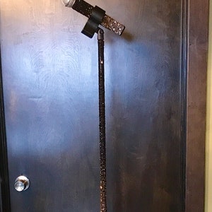 Puede incluir: Un micrófono negro sobre un soporte, ambos adornados con brillantes detalles negros y plateados. El micrófono está inclinado hacia el espectador, y el soporte es alto y delgado. El fondo es una puerta de madera oscura.