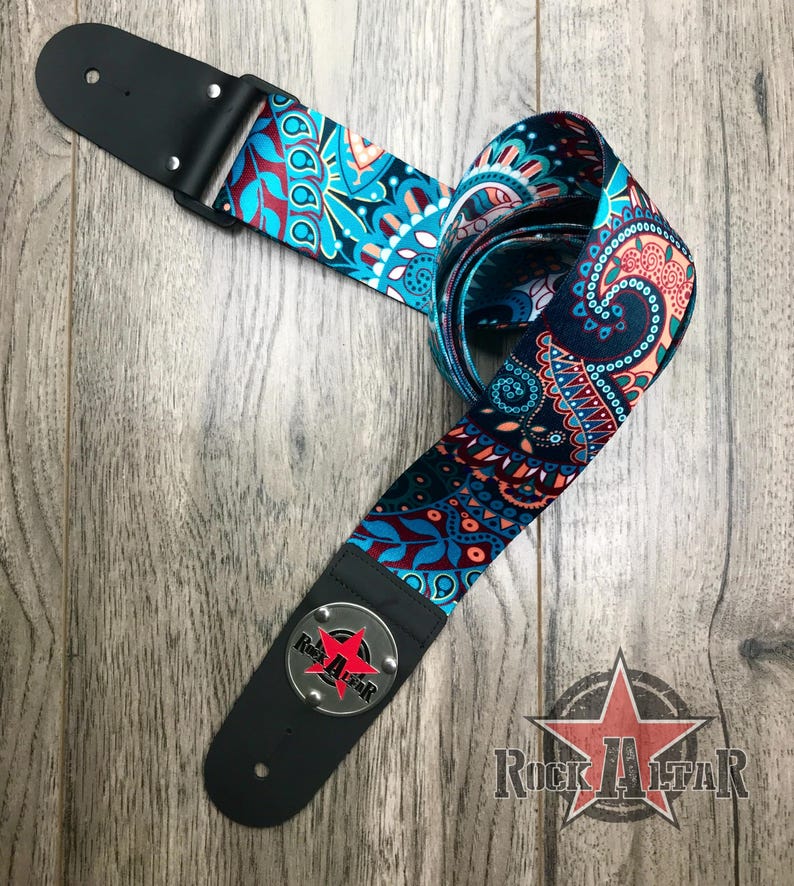 May include: A guitar strap featuring a vibrant paisley pattern in shades of teal, dark blue, and coral orange. The strap includes black leather ends with silver rivets and a circular metal emblem displaying a red star and the text Rock Altar.