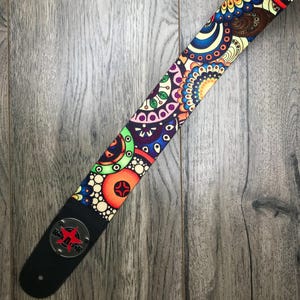 May include: A guitar strap featuring a vibrant, multi-colored circular pattern with shades of orange, blue, green, and purple. The strap includes a black leather end piece with a circular silver metal emblem displaying a red star and the text Rock Gear.