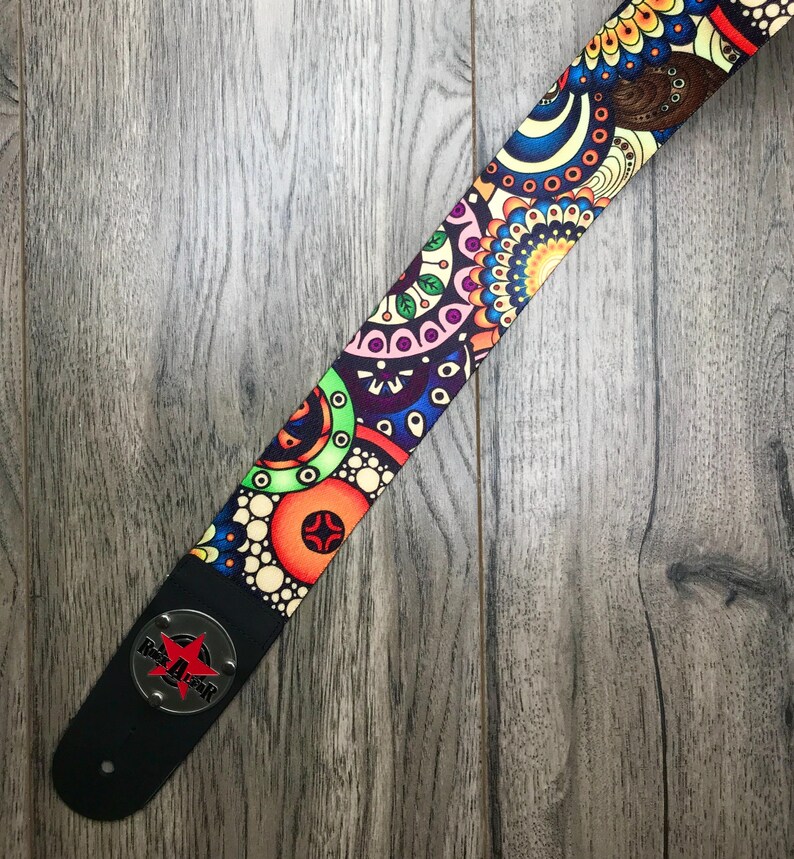 May include: A guitar strap featuring a vibrant, multi-colored circular pattern with shades of orange, blue, green, and purple. The strap includes a black leather end piece with a circular silver metal emblem displaying a red star and the text Rock Gear.