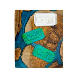 May include: Three cookie cutters on a wooden cutting board with a blue and brown epoxy resin design. The cutters are shaped like a can of Diet Coke. One cutter is white and the other two are green.