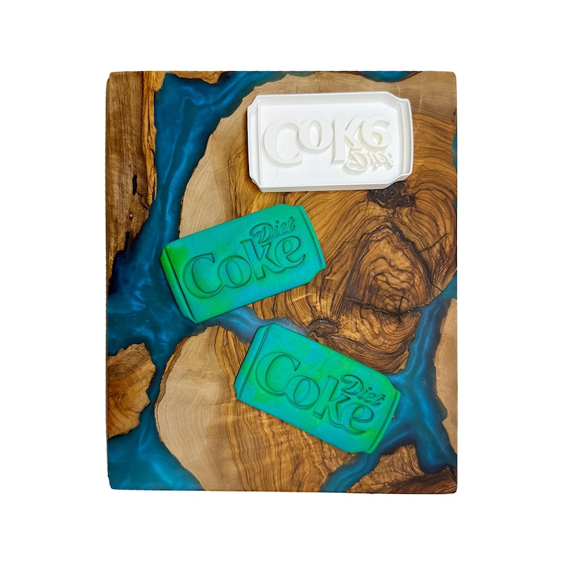 May include: Three cookie cutters on a wooden cutting board with a blue and brown epoxy resin design. The cutters are shaped like a can of Diet Coke. One cutter is white and the other two are green.