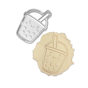 May include: A silver-colored cookie cutter in the shape of a smiling bubble tea cup with boba pearls. Next to it is a piece of dough with the bubble tea design pressed into it, showing the impression of the cutter.