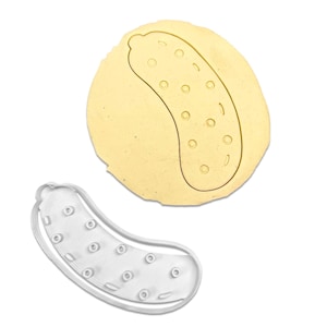 May include: A pickle-shaped cookie cutter next to a piece of dough with a pickle imprint. The cookie cutter is made of silver-colored metal and has a detailed outline of a pickle with circular indentations. The dough is light yellow.