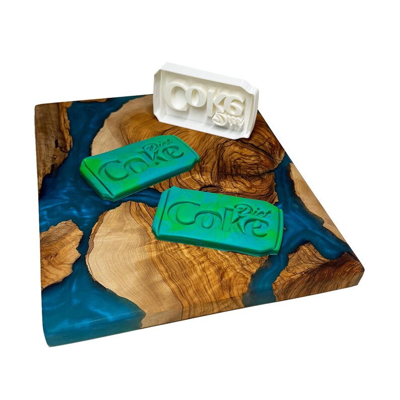 May include: A wooden cutting board with a blue epoxy resin inlay. There are three silicone molds on the board, two green and one white. The molds are shaped like a can of Diet Coke and have the words "Diet Coke" embossed on them.