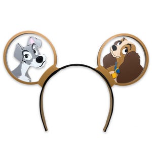 May include: A pair of brown mouse ears with a cutout of Lady and the Tramp. Lady is on the right ear and Tramp is on the left ear.