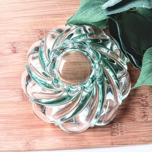 May include: A clear glass decorative bowl with a spiral design and a wooden-like center. The bowl has a light teal tint and sits on a wooden surface. Green leaves are in the background.