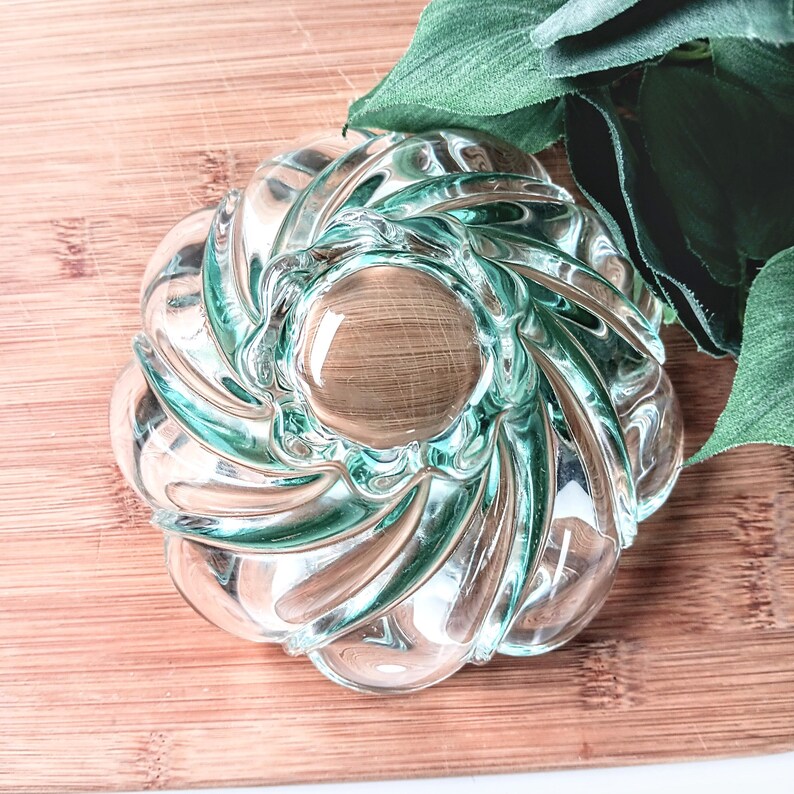 May include: A clear glass decorative bowl with a spiral design and a wooden-like center. The bowl has a light teal tint and sits on a wooden surface. Green leaves are in the background.