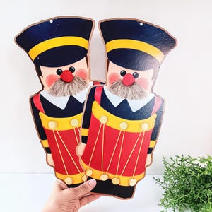 May include: Two wooden nutcracker ornaments with black hats, red and white faces, and yellow and red accents. The nutcrackers are holding drums.