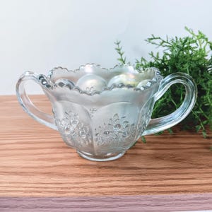 May include: A clear glass sugar bowl with two handles and an iridescent finish. The bowl has a scalloped rim and is decorated with an embossed floral pattern.