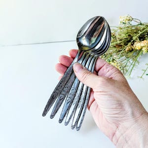 May include: A set of five silver-toned metal spoons featuring a detailed floral pattern embossed along the length of each handle. The spoons have a polished finish and are arranged in a fan shape, showcasing the intricate botanical engravings.