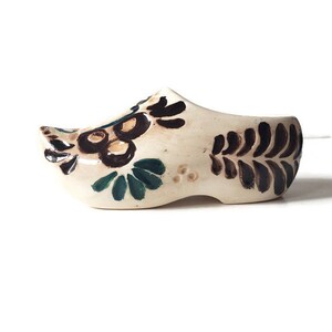 May include: A small, off-white ceramic clog with hand-painted floral designs in brown and dark green. The decorative shoe features a rounded toe and a curved heel, showcasing a traditional Dutch design.