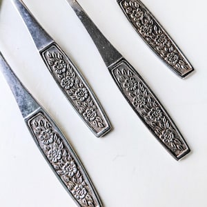 May include: Close-up of four vintage silver forks. The handles feature an embossed floral design. The forks are arranged diagonally against a white background, showcasing their intricate details and aged appearance. The silverware is likely part of a dining set.