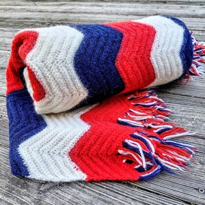 May include: A rolled-up, chevron-patterned blanket in red, white, and blue. The blanket has a fringed edge with matching colors. The design features a zig-zag pattern, with the colors arranged in stripes. The blanket is made of a soft, textured material.