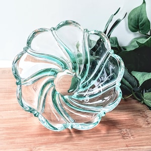 May include: A clear glass bowl with a swirling, flower-like design. The bowl has a light teal tint and sits on a wooden surface. The edges of the bowl are scalloped, and the overall shape is circular.