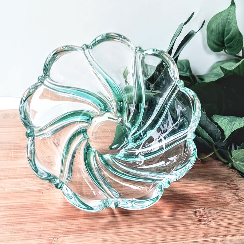May include: A clear glass bowl with a swirling, flower-like design. The bowl has a light teal tint and sits on a wooden surface. The edges of the bowl are scalloped, and the overall shape is circular.