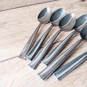May include: A collection of six stainless steel spoons. Each spoon has an oval bowl and a long, angular handle. The spoons are arranged diagonally on a light-coloured wooden surface.