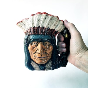 May include: A ceramic pitcher shaped like an Native American chief wearing a feathered headdress. The pitcher is painted in shades of brown, red, white, and blue.