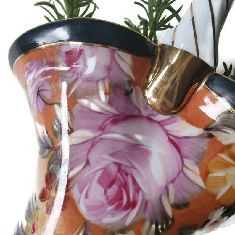 May include: A white ceramic planter with a gold rim and a floral pattern. The planter has a handle and is shaped like a teapot. The planter is filled with green plants.