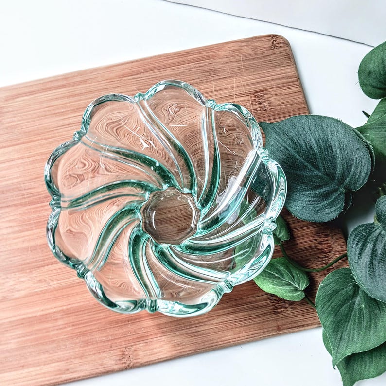 May include: A clear glass bowl with a swirling, petal-like design. The bowl has a central, circular base and a scalloped rim. The glass has a slight green tint. The bowl is on a wooden cutting board.