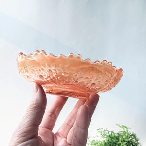 May include: A small, vintage, peach-colored glass bowl with a scalloped edge and a floral design. The bowl is made of iridescent glass and has a delicate, feminine look.