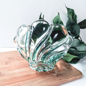 May include: A clear glass decorative bowl with a shell-like design. The bowl has a wavy, textured surface and is placed on a wooden cutting board. Green leaves are visible in the background.