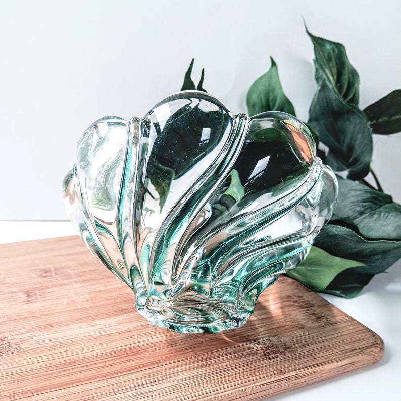 May include: A clear glass decorative bowl with a shell-like design. The bowl has a wavy, textured surface and is placed on a wooden cutting board. Green leaves are visible in the background.
