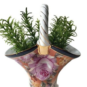May include: A white porcelain basket with a twisted handle, decorated with a floral pattern in shades of pink, orange, and yellow. The basket has a blue and gold trim and is filled with green sprigs.