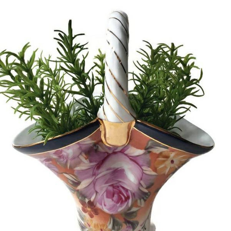 May include: A white porcelain basket with a twisted handle, decorated with a floral pattern in shades of pink, orange, and yellow. The basket has a blue and gold trim and is filled with green sprigs.