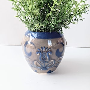 May include: A small, blue and brown ceramic planter with a floral design. The planter is filled with green leafy plants.