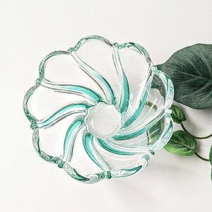 May include: A clear glass bowl with a flower-like design. The bowl features a central white circle and teal-colored swirls radiating outwards. The rim has a scalloped edge. A green leaf and vine are visible in the background.