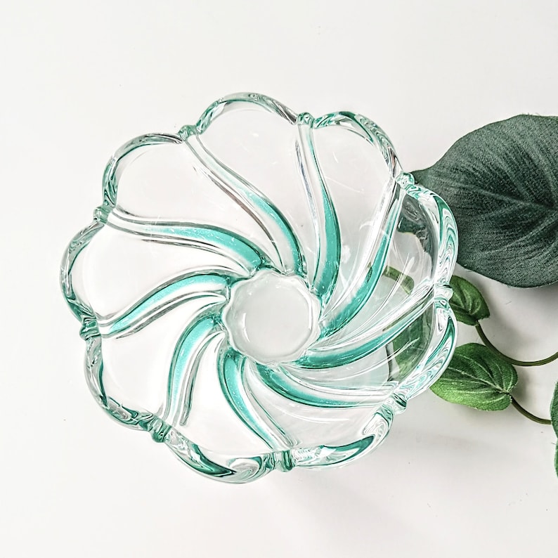 May include: A clear glass bowl with a flower-like design. The bowl features a central white circle and teal-colored swirls radiating outwards. The rim has a scalloped edge. A green leaf and vine are visible in the background.